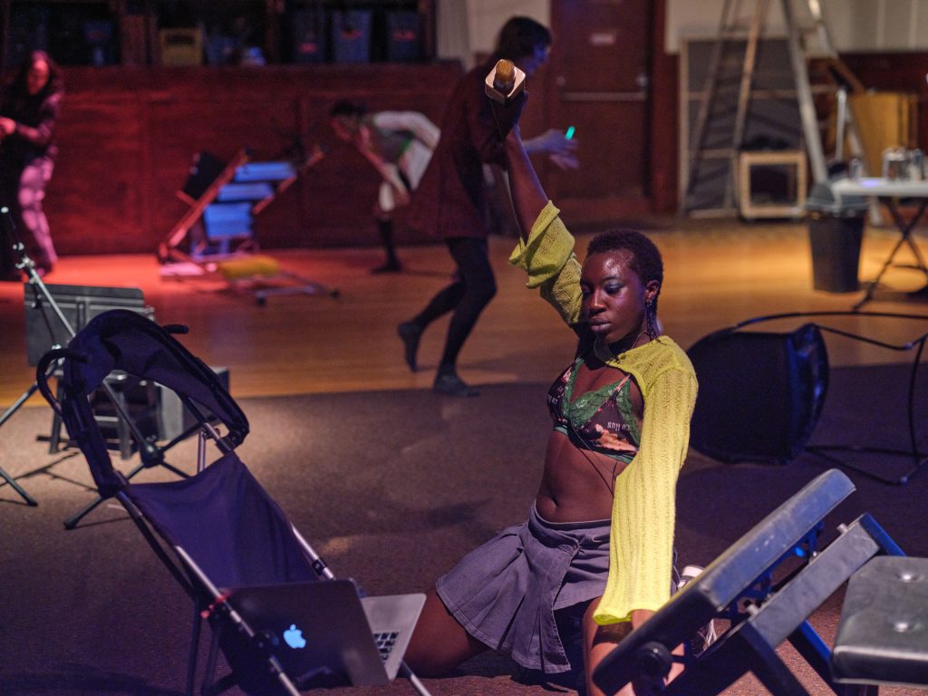 Samra squats on her knees waving a karaoke microphone above her head. She reads from a laptop perched in a folding baby stroller in front of her. 