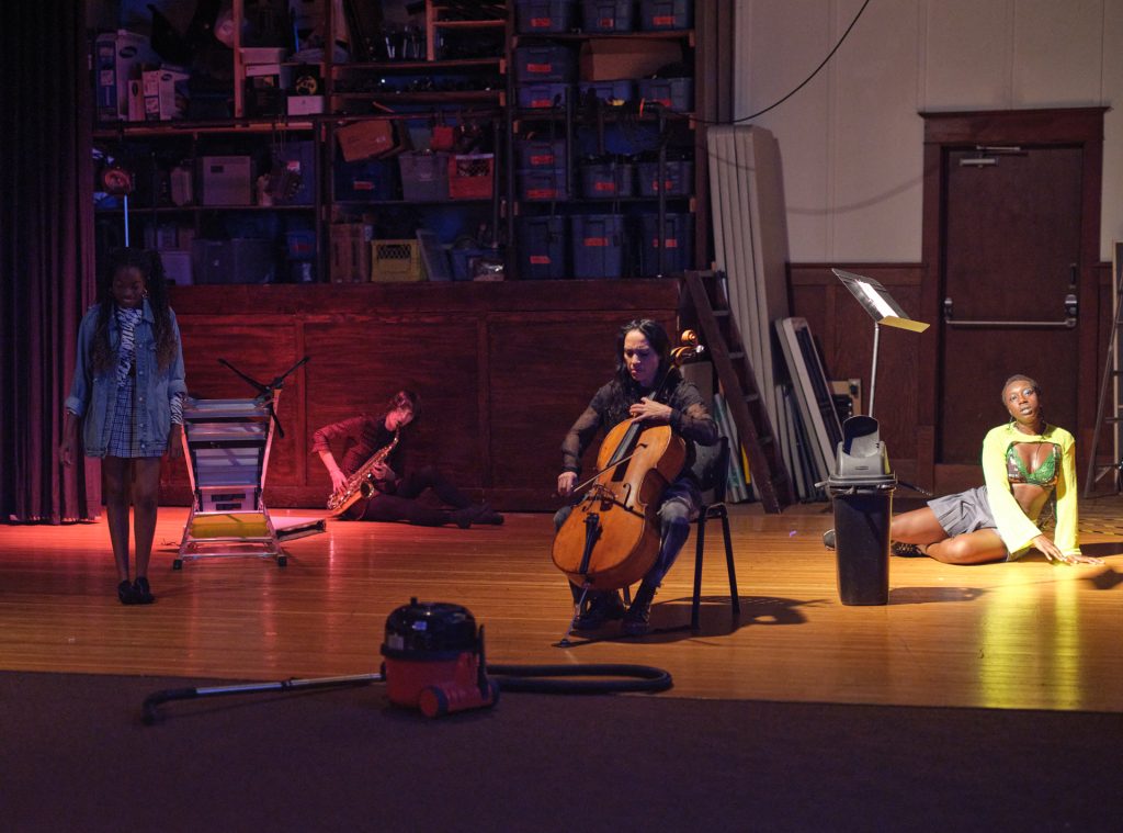 Four figures scattered amongst music and maintenance objects. A woman with braids smiles at her feet, behind her a person playing a saxophone sits on the floor, a cellist sits on a chair, Samra sits on the floor gazing into the distance. 