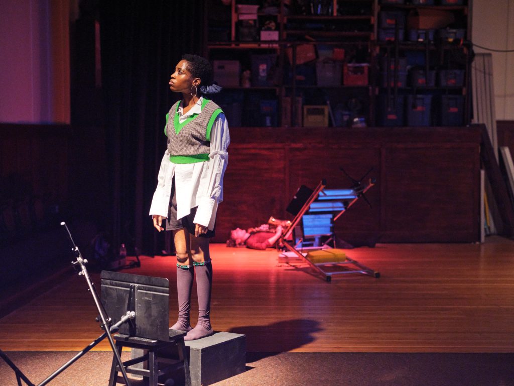 A woman wearing knee socks, an oversized white button up shirt and mini cropped grey and green sweater vest stands on a box and gazes upwards. A fallen music stand lays on a stool at her feet. 