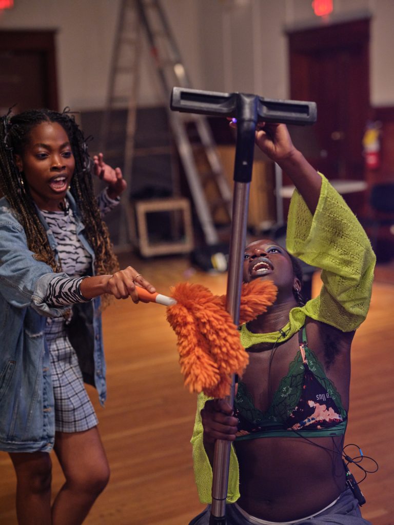 Samra holds up a vacuume cleaner as a singer with braided pigtails dusts it with a fluffy orange duster. Both are singing mid action. 