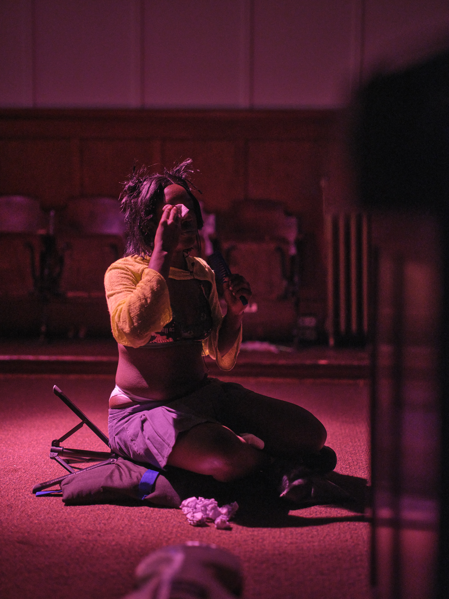 A woman wearing a black wig sits on the ground with her legs tucked under wiping makeup off her face in a pool of raking pink light. High rise bottoms peak out of a low rise grey skirt, a green bathing suit top shows under a yellow knit extra cropped sweater. Crumpled paper litters the ground around her.