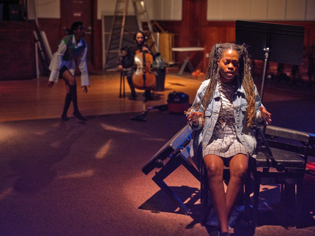 A woman with long brown braids in pigtails sits on a pile of piano benches and music stands, her hands are lifted at her sides conducting the song she is singing.
