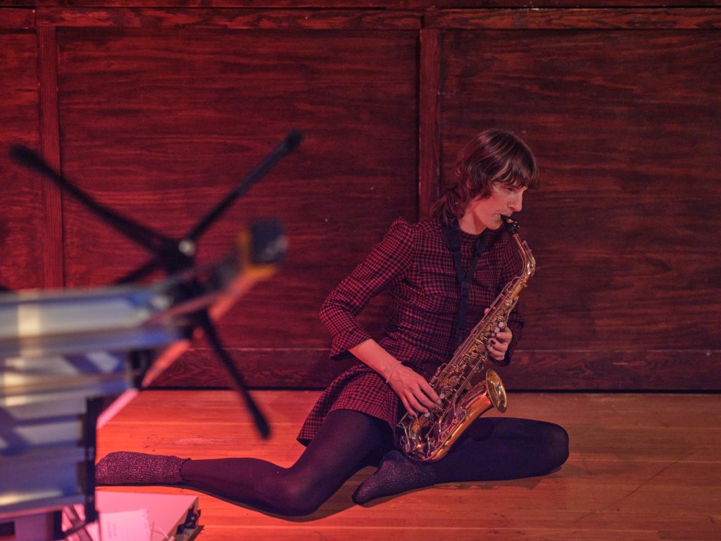 A person in a red and black tartan dress sits on the floor playing saxophone.