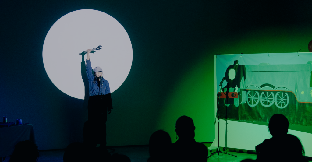 Megan, wearing coveralls and a train conductors cap, stands in a spotlight and speaks into a microphone while  holding up a wrench in a gesture of workers unification.