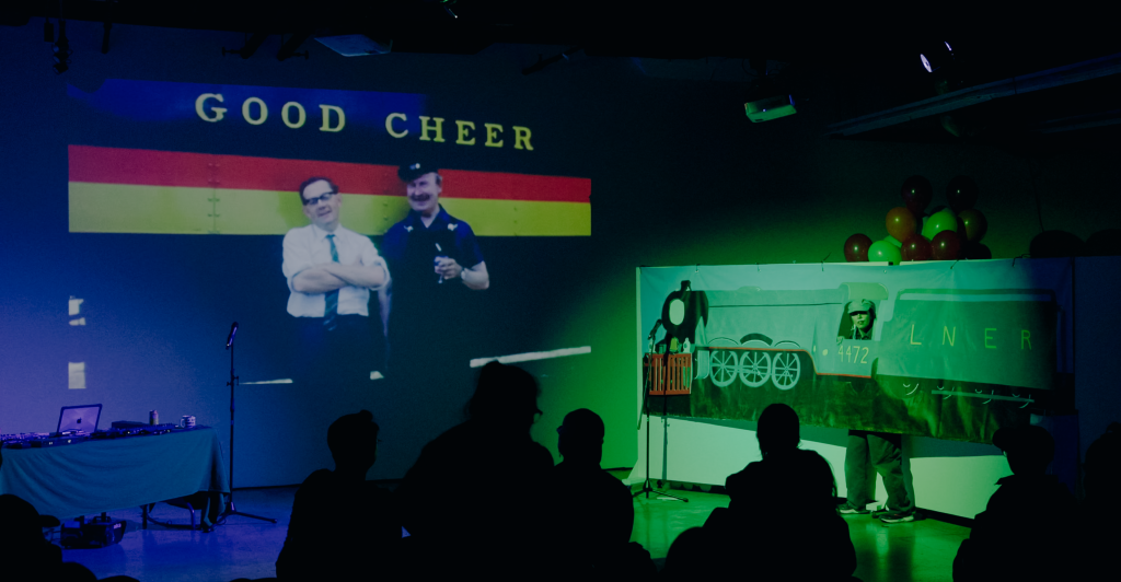 Megan poke out from behind the train painted vinyl banner in a conductors cap. In a projection, two men lean against the side of a train, the words: Good Cheer, are written above their heads. 