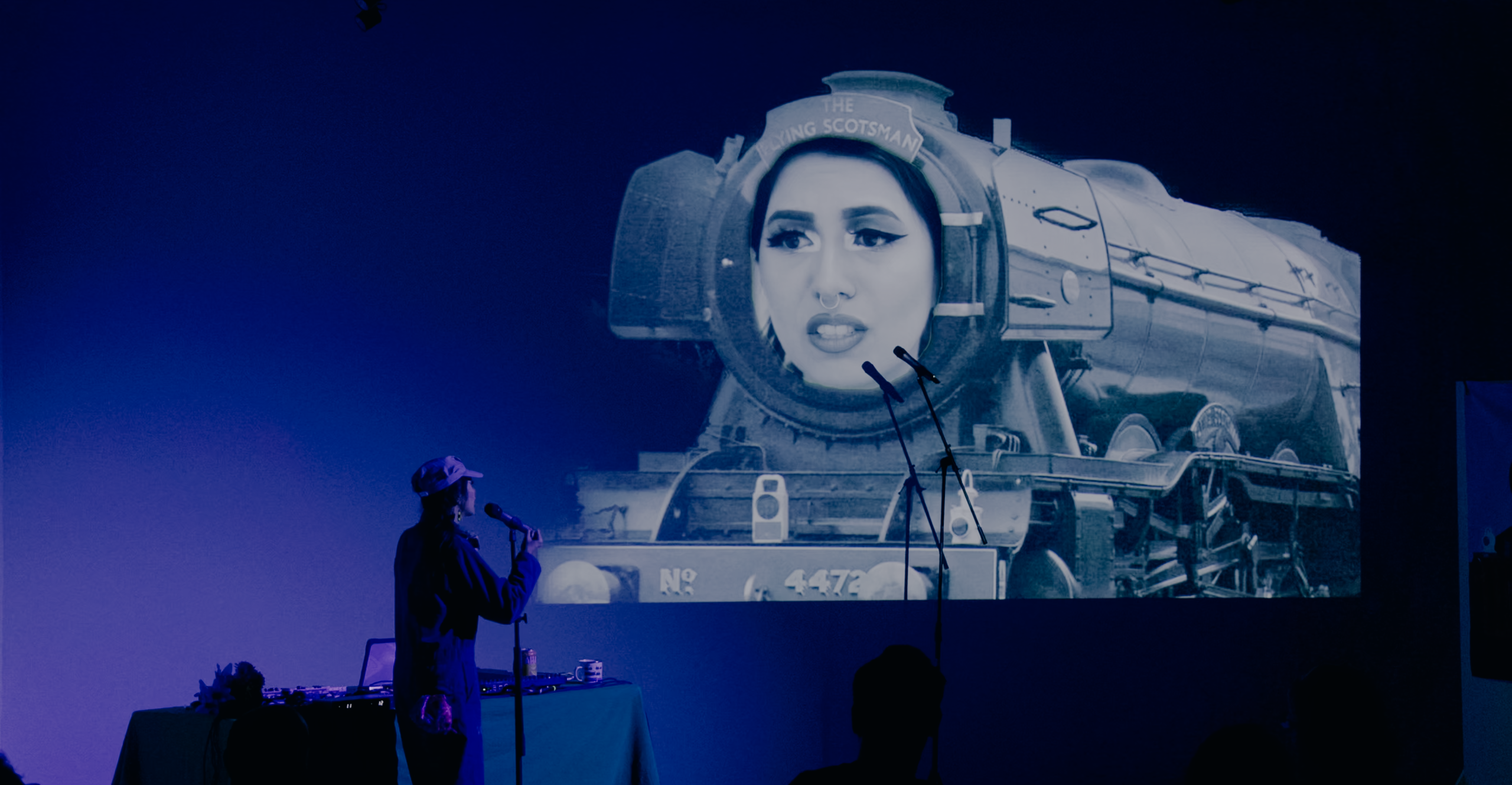 Megan, wearing blue coveralls and a train conductors cap sings toward a projection of the Flying Scotsman train with their face superimposed on top. The pair sing to each other through microphones on stands.