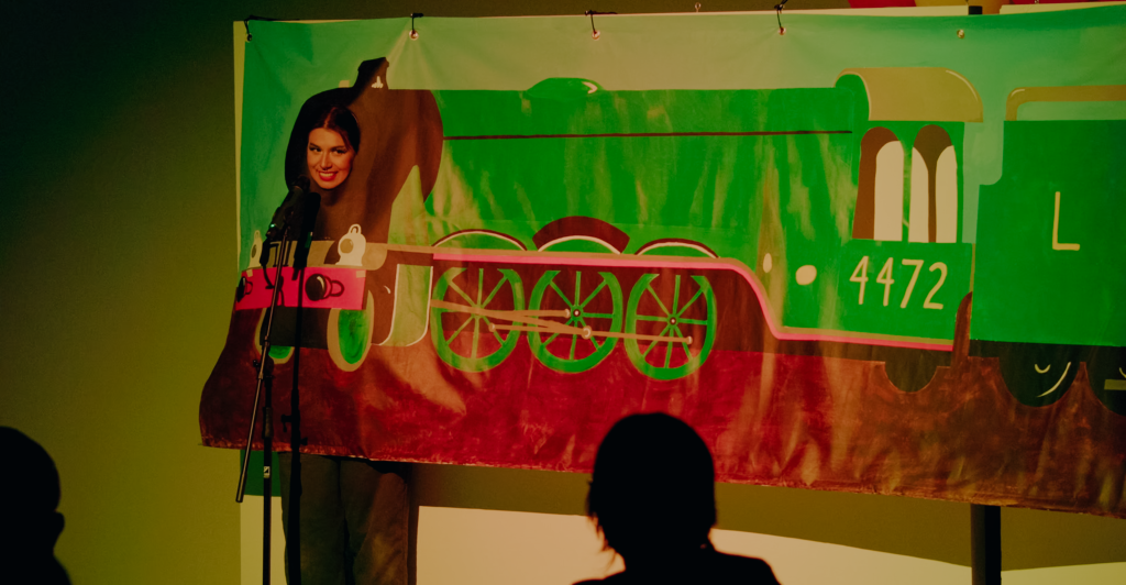 Megan sticks their smiling face through a vinyl banner painted with a green locomotive 4472 engine and speaks into a microphone. 