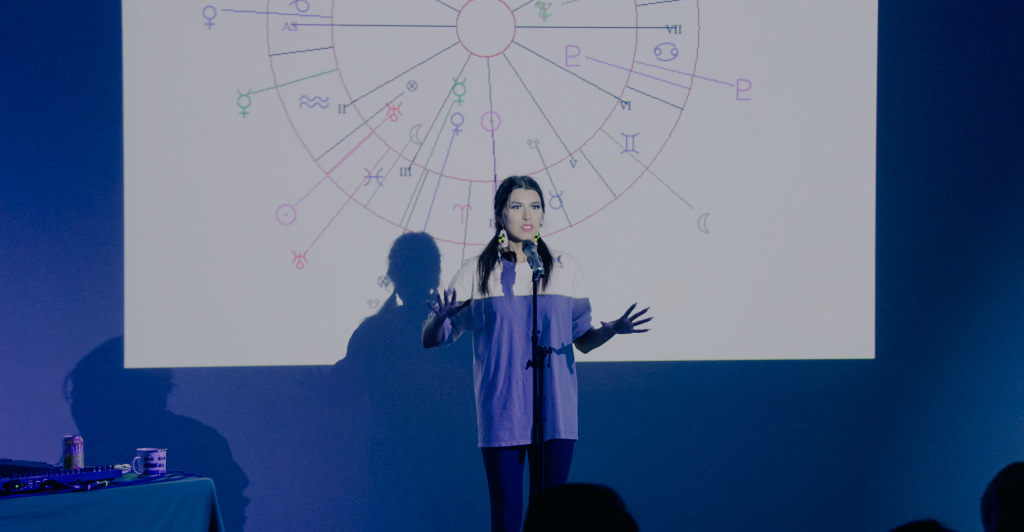 Megan, wearing a white t-shirt, gesticulates while speaking to the audience in front of a projection of an astrological chart. 