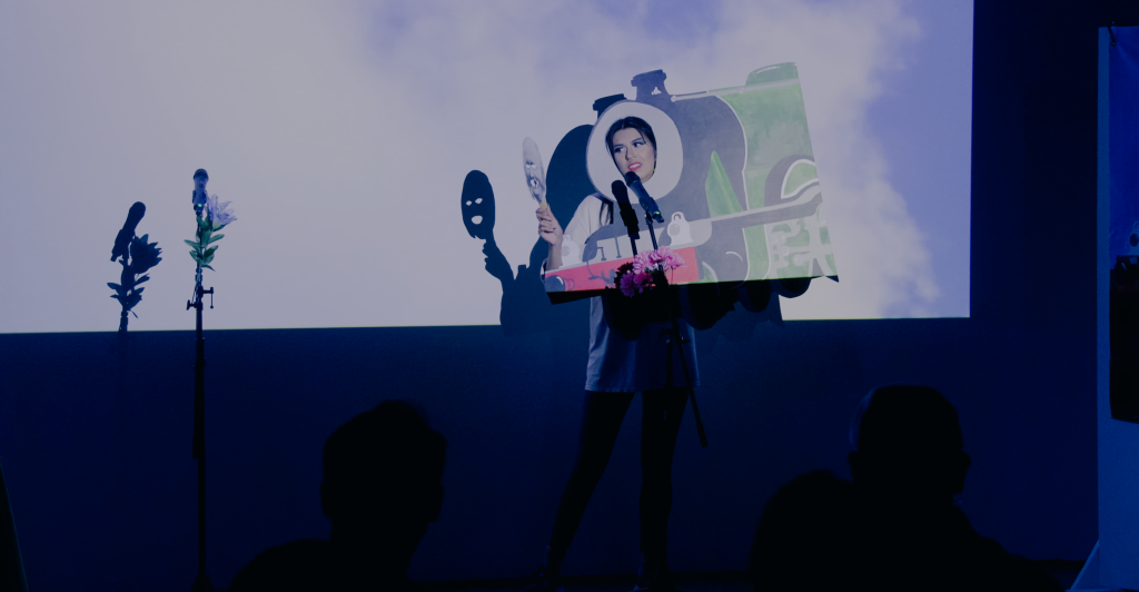 Megan's head sticks through a partial train engine cut out mask as they sing to a paper mask of Alan Pegler.