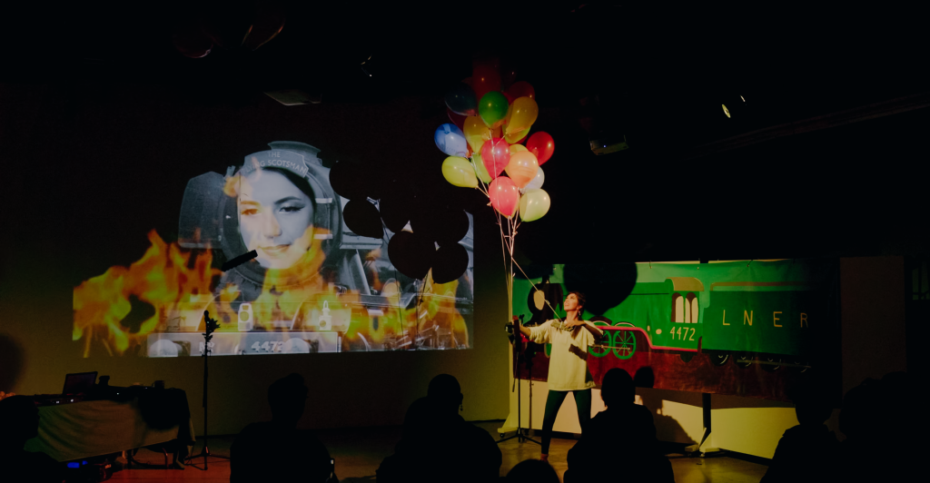 Megan releases a tangle of toy train engine's tied to floating balloons. In a projection, the Flying Scotsman train smiles as fake flames engulf the bottom of the screen.