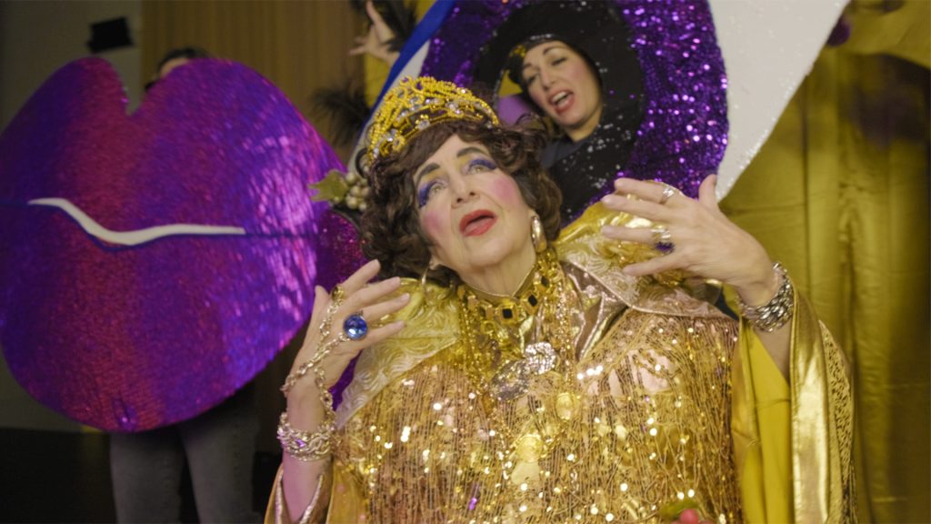 An older woman with bold makeup sings theatrically while dressed in layers of gold sequins and sparkles. 