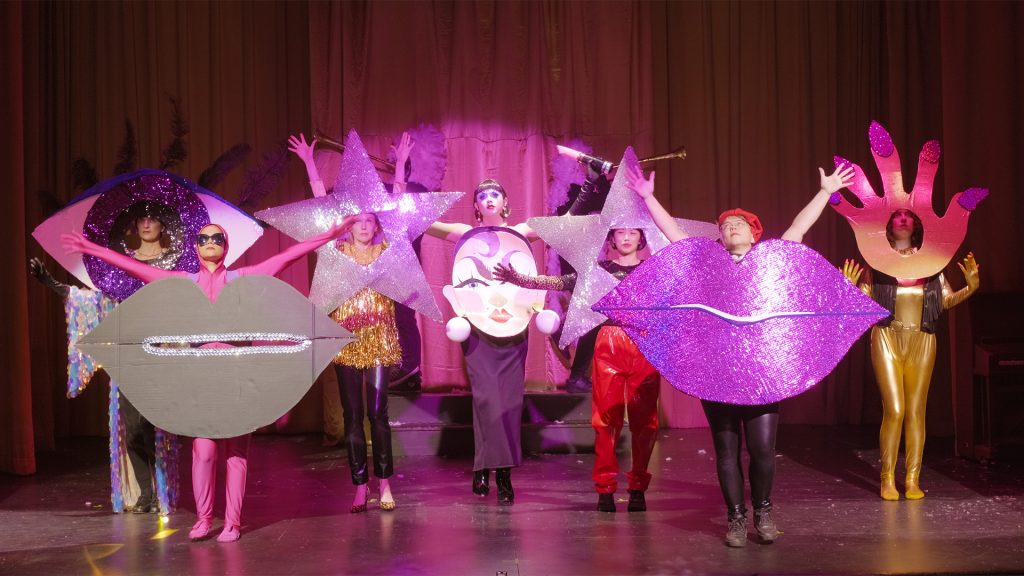 A cast of characters wearing sparkling cardboard cut out costumes of lips, eyes and hands stand like stars on stage bathed in pink lights.