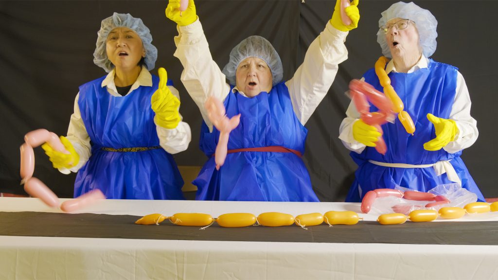 Three women wearing blue aprons, yellow gloves and hairnets toss plastic sausages around while singing. 