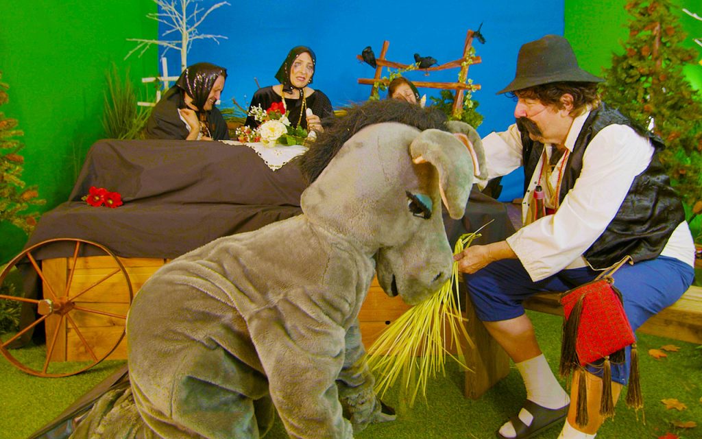 A man with a large fake moustache feeds straw to an actor in a donkey costume as three women in mourning clothes cry over a casket.