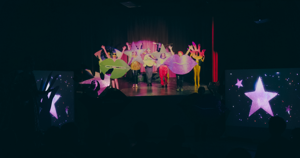 In a dark room three projector screens illuminate shadowy figures. Pink stars shine as a cast of characters wearing sparkling cardboard cut out costumes of lips, eyes and hands dance in unison.