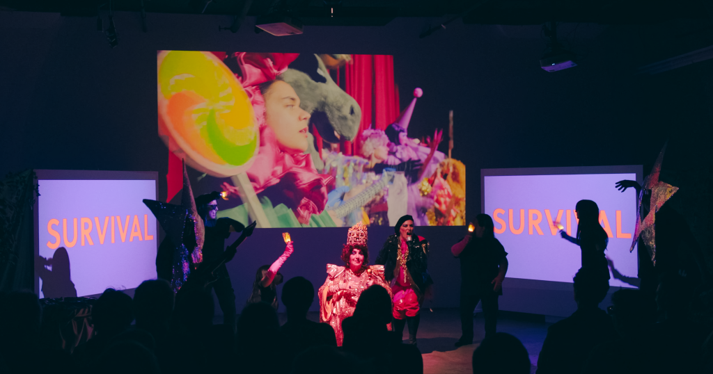 A cast of characters dance framed by three projector screens. The word Survival is written on the screens on either side while the central screen plays a video of actors dressed as animals and flowers while on stage. 
