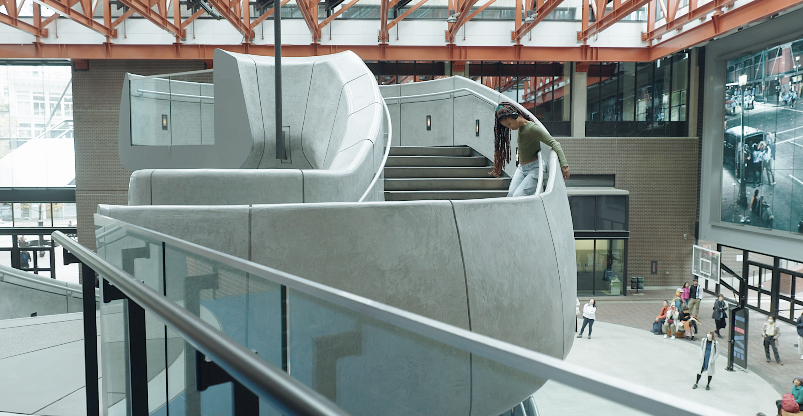 Jessica, wearing a green shirt and jeans, snakes her way down a sinuous concrete stairwell. Large over ear headphones frame her face and dreadlocks tied up in a high pony tail.