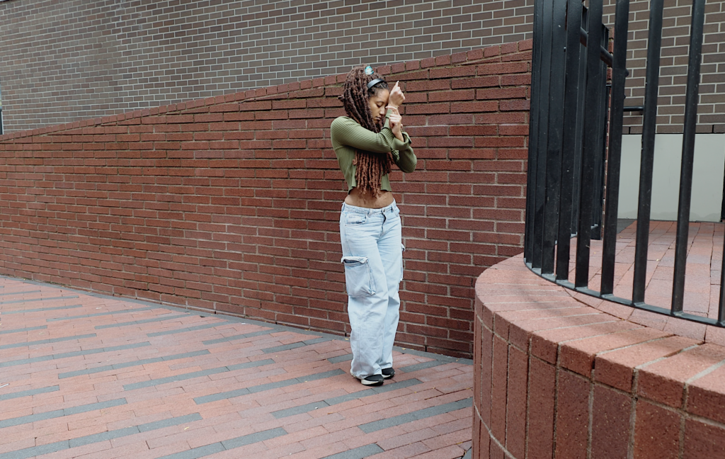 Jessica wraps her arms up close to her face as she shuffles beside a brick wall, listening to the music in her black headphones. 