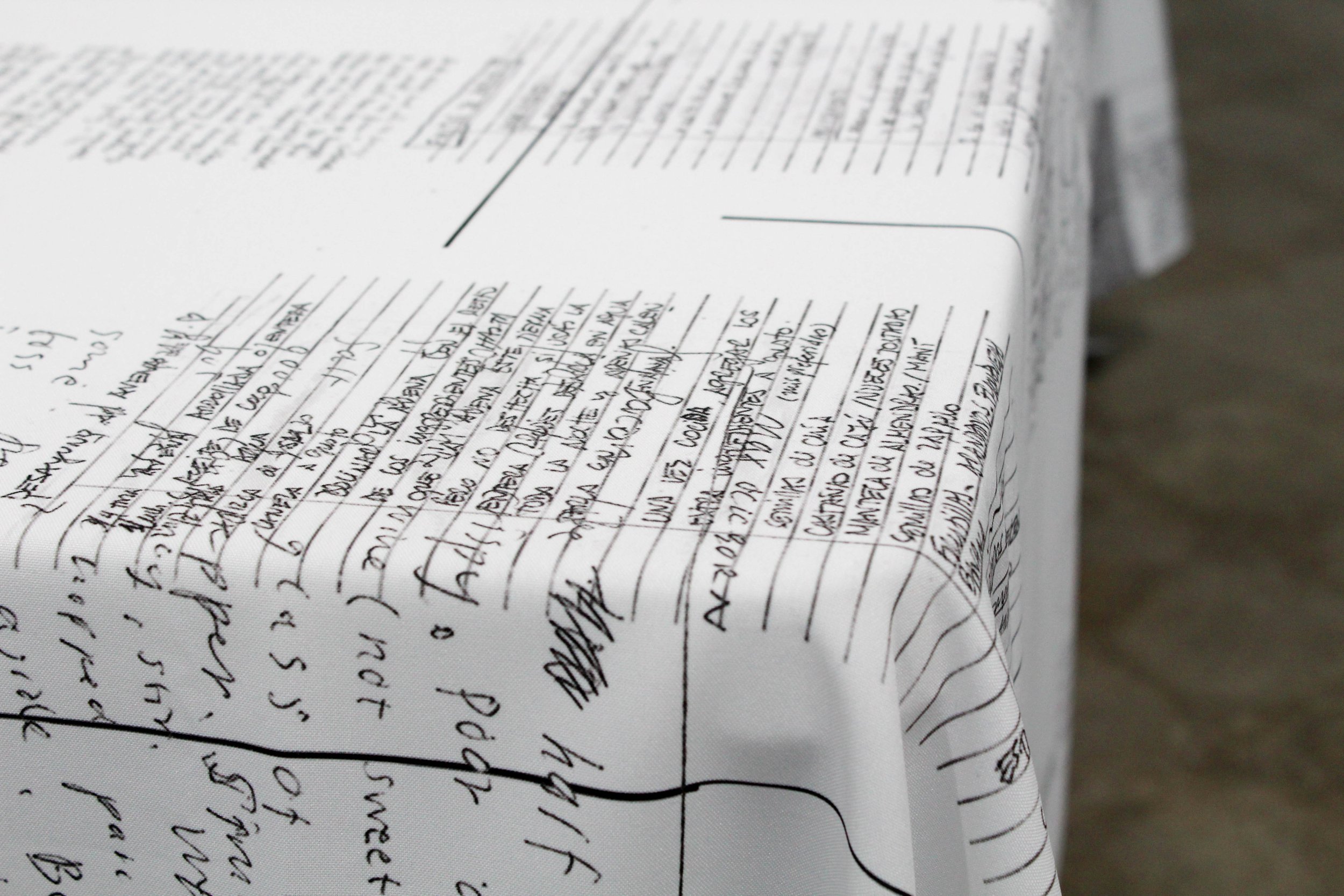 A long table stretches out from the corner in focus. It is lined with a white table cloth inscribed with hand written recipes and scribbled notations.
