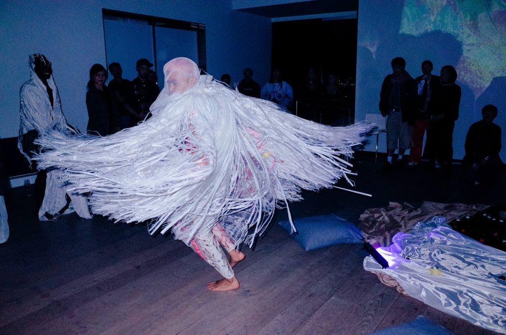 A dancer twirls in a sea of white tassels, like a jellyfish made of silk and ribbon they step mid glide thorough the audience.