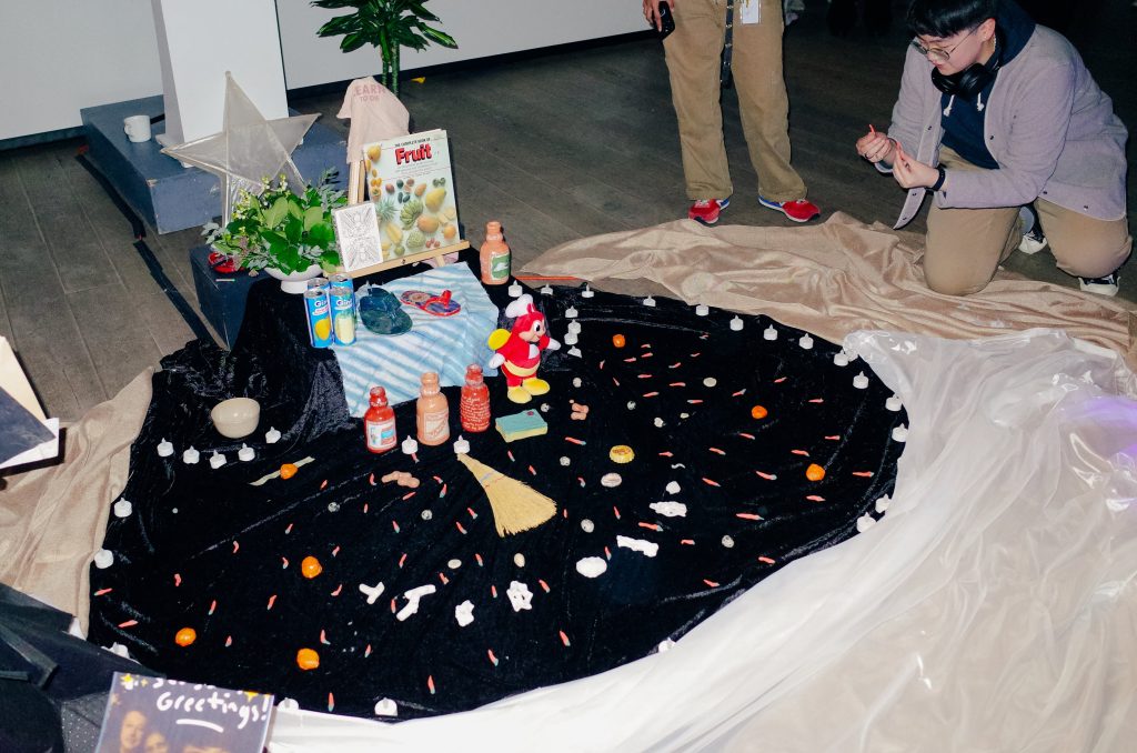 An circular alter on black velvet: rings of candies, fruit juice, foliage and electric tea lights are being added to by kneeling participants. 