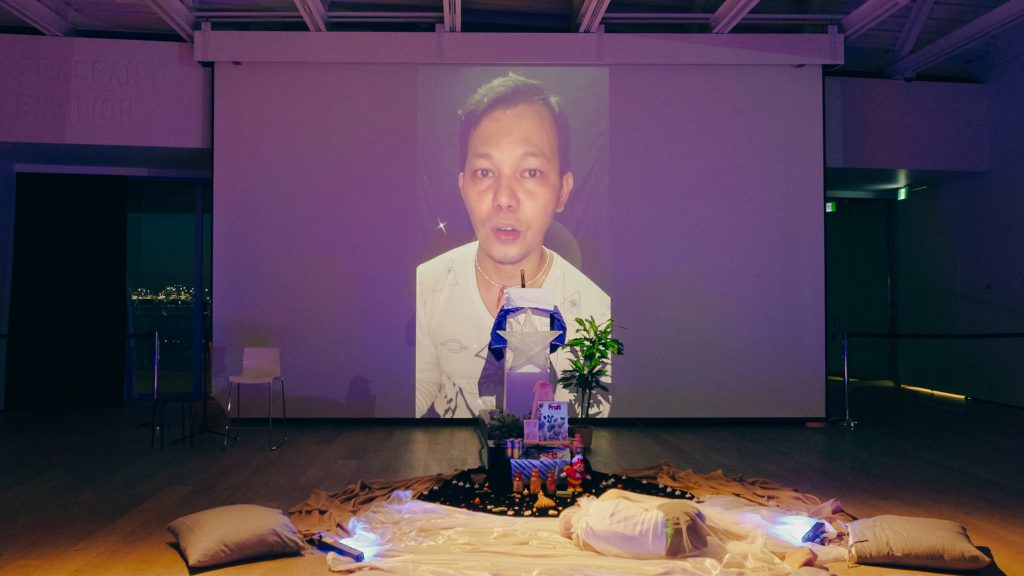 A body curls under a sheer cloth, lying on a circle of fabrics surrounding an alter of food and object offerings. A video of Zeus Bascon speaking to the camera is projected behind this scene. 