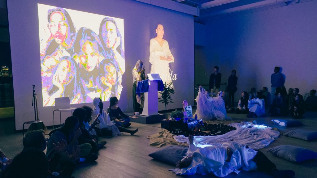 A shawled Jordan Baylon speaks from an alter. Multiple multicoloured faces are projected behind an alter encircled by viewers, giving the room a blue glow. 