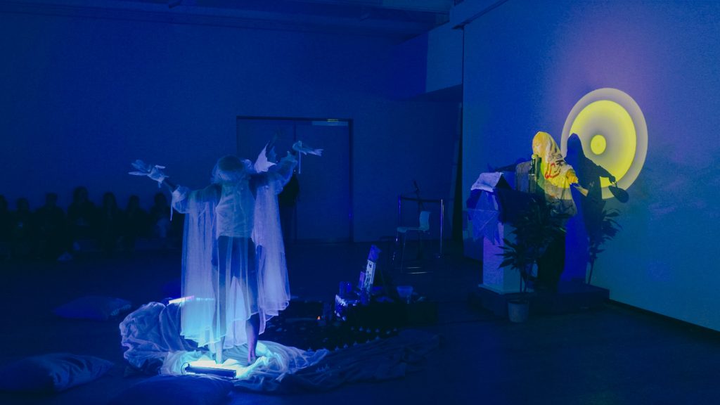 Jordan Baylon stands at an alter in a halo of yellow light. Two dancers cloaked in sheer white cloths stretch in the centre of the circle. 