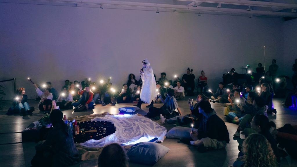 A performer cloaked in white silk weaves through the audience seated on the floor around an alter of fabric. Orbs of light dot the room from flashlights and phones. 