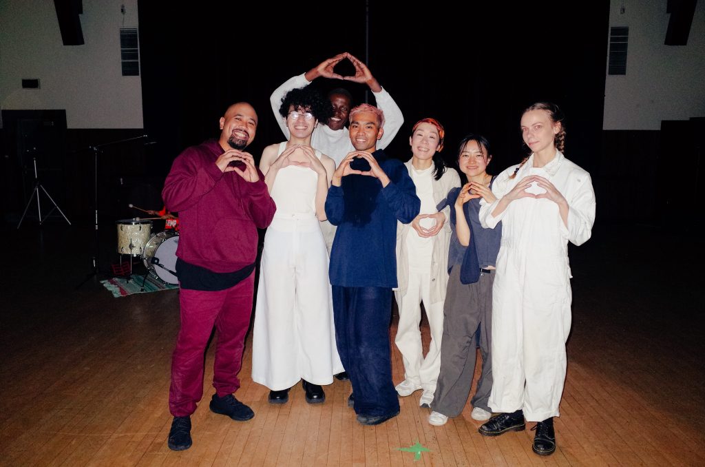 A group of seven people wearing earthy tones and white baggy garments pose smiling in a line, their hands are raised at their chests forming the shape of a circle.