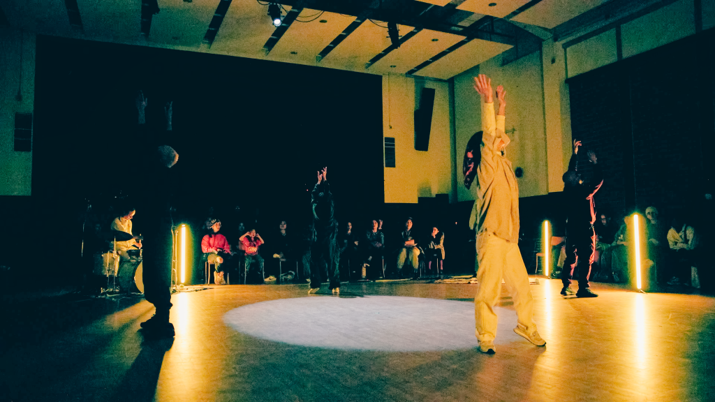 Four dancers face the audience in a circle, they stretch their arms to the sky, their bodies dappled in the warm lights surrounding them.