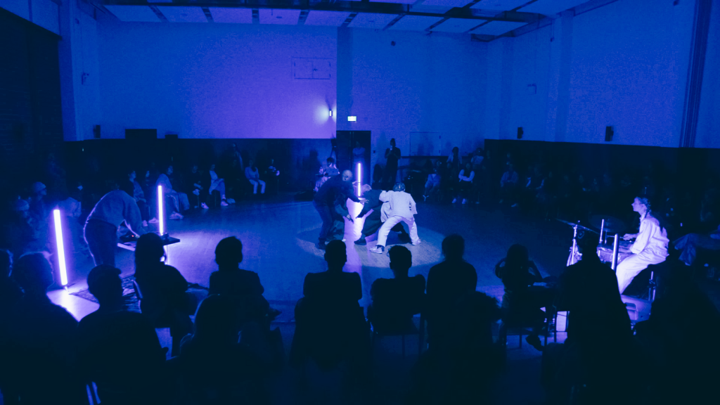 A seated audience encircles a group of dancers in a darkened room. Blue lights encapsulate the figures as they grove close to each other, close to the ground.