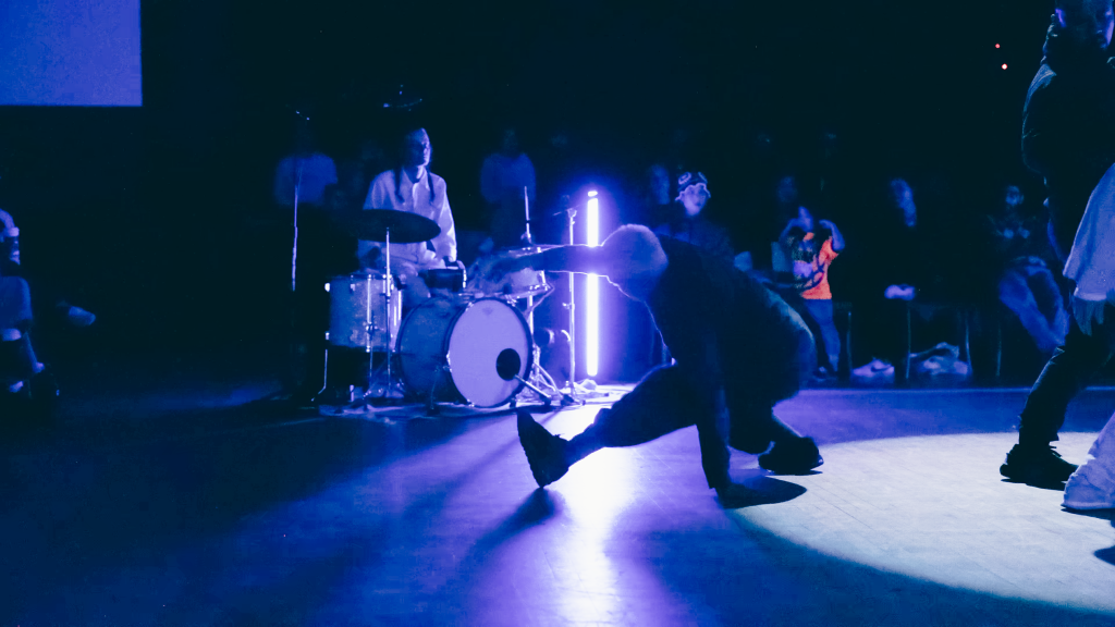 A dancer slide lunges across the floor, using their arm as a pivot point. Blue lights encase them and a drummer dressed in white in the background.