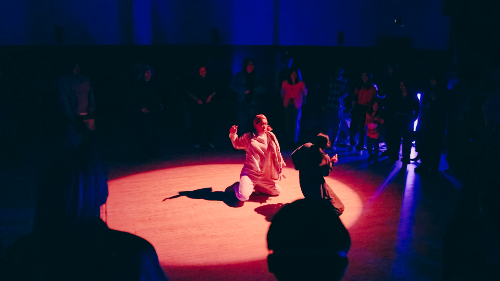 A dancer in white baggy clothes crouches in the center of a spotlight, a slide on knees travels them towards another dancer.