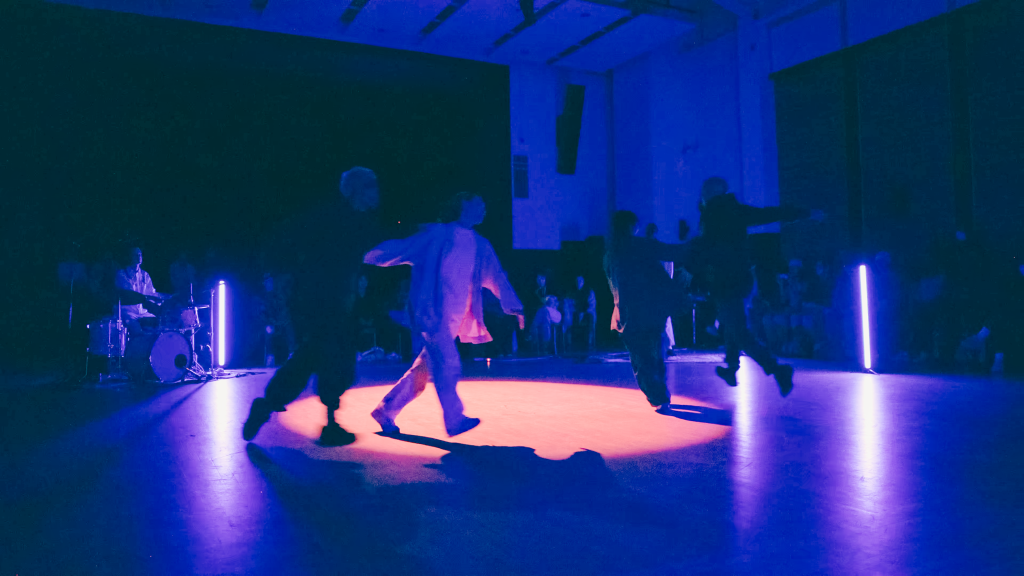 A group of dancers run in a circle around a spotlight, the light cuts through the blue darkness of the room.