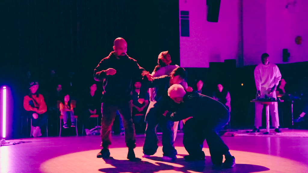 A group of dancers mingle in the center of a spotlight. Pink lights cast sunset shadows around their shifting bodies.