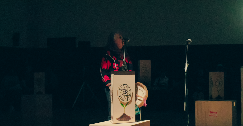 A woman in a black sweater with large red shapes sings into a microphone, a hide drum hangs in her hand by her side.