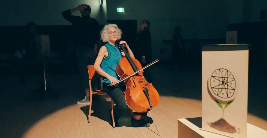 A grey haired women sits in a spotlight playing a cello. A triangular light box with a circular line drawing rests in the foreground.