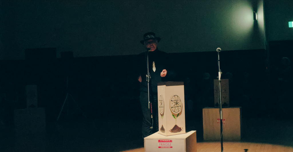 Michelle, wearing a black turtleneck, a fedora hat and a large bone necklace, speaks into a microphone. A wooden light box rests in the foreground.