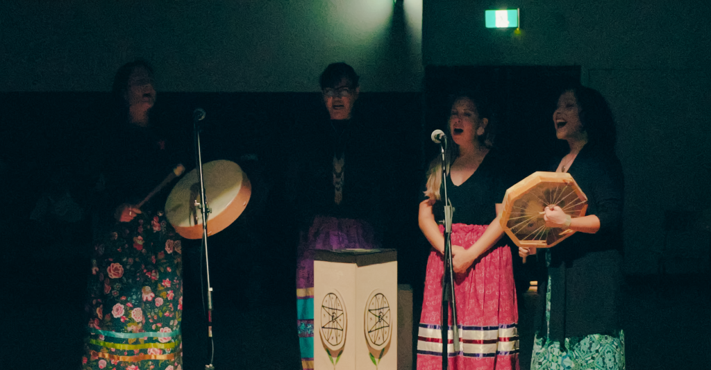 Four women wearing colourful floral and ribbon skirts sing into microphones in a dimly lit room. Two of the women are playing hide drums.