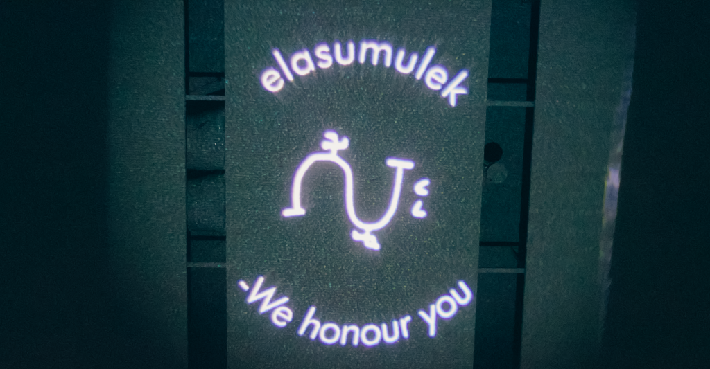 A light projection on the celling casts a spotlight in a darkened room. A squiggly line is surrounded by the words, elasumulek - we honour you.