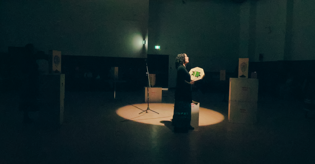 A woman weaves around wooden light boxes while pacing the parameter of a spotlight. She is playing a hide drum and singing.