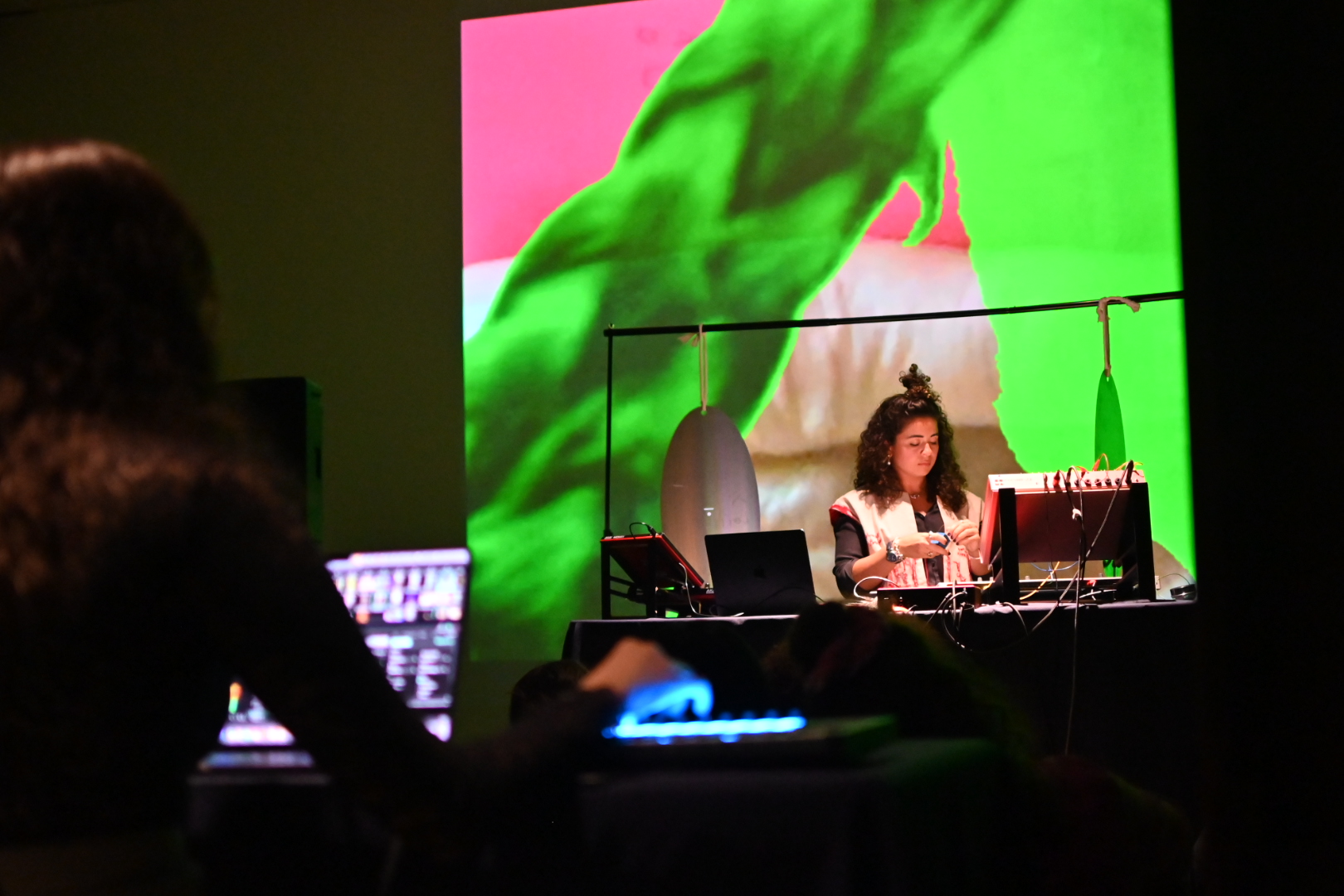 Looking over Saghi's shoulder we see their laptop and VJ set up, they are looking across at Kimia who is setting up her audio equipment at a table in front of Saghi's station. Abstract forms in greens and pinks dance in a video projection behind both artists.