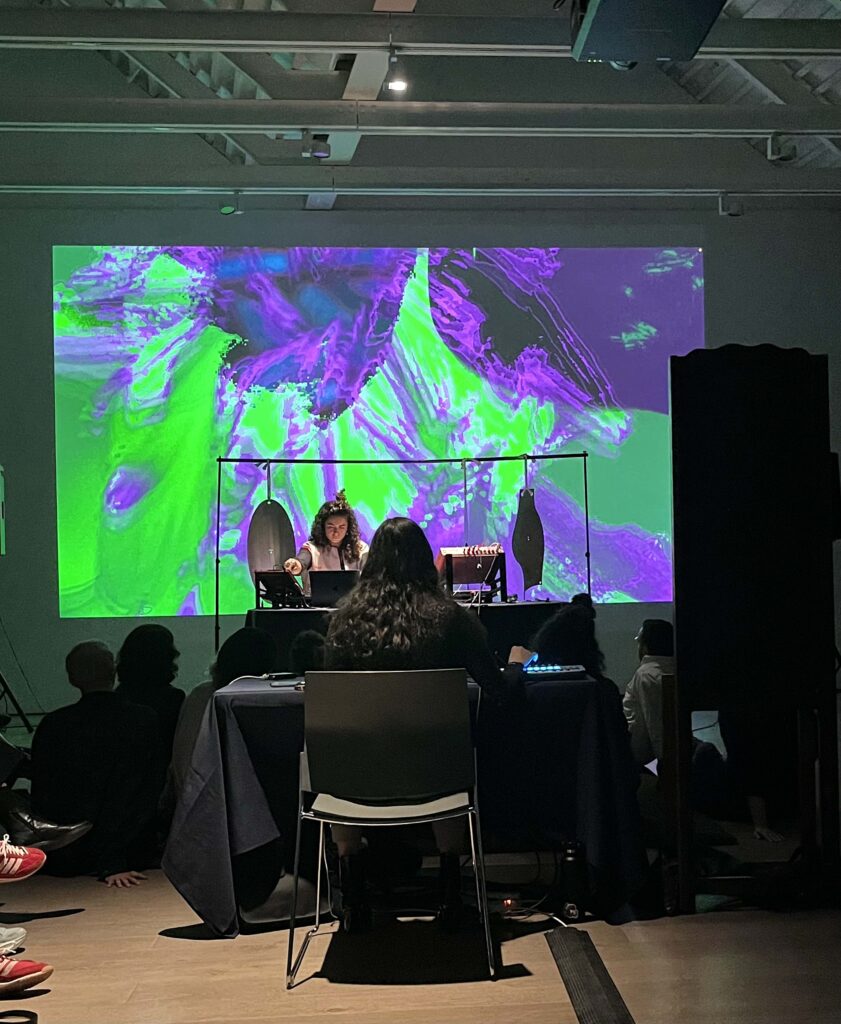 Saghi sits at a table, their long black hair running down their back facing the camera. Across from them, Kimia is tweaking with her audio equipment at another table. Saghi is projecting a green and purple image of layered flowers on to the back wall.