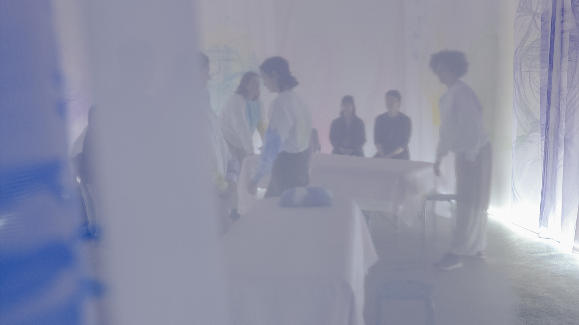 A group of people move like ghosts, blurred behind a gauzy curtain. Artists in tie-dyed shirts move massage tables covered in blankets as the audience watches.