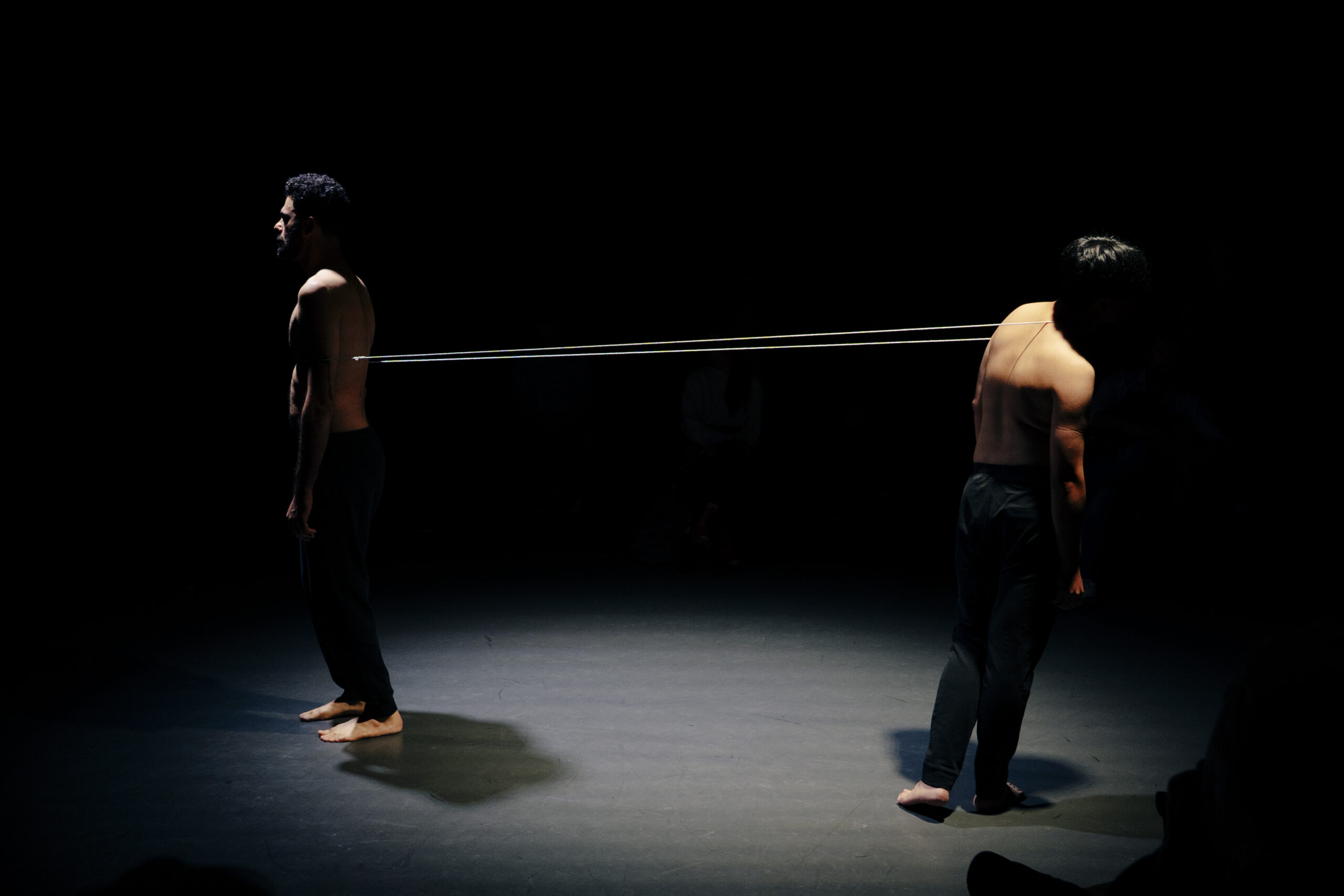 Under a spotlight in a darkened room, two shirtless bodies tethered by a small rope rotate in tension with one another. The rope digs into Aryo's chest and crosses upward to Sidi's neck which is tilted away. Both wear black pants and have bare feet.