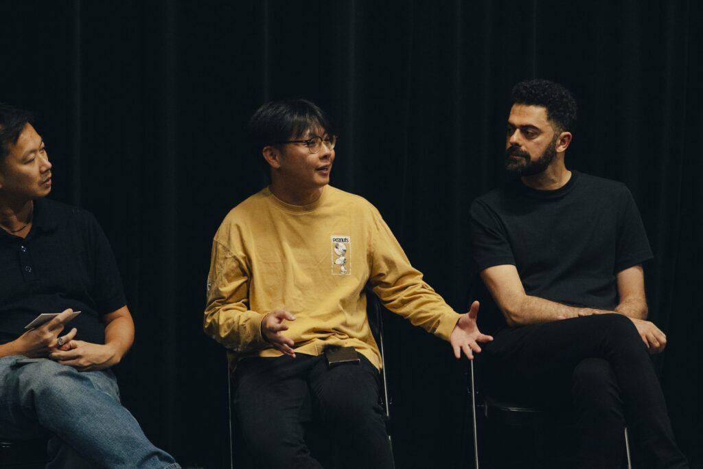 Derrick, Sidi and Aryo sit in a row of chairs. Derrick and Aryo look at Sidi who is gesticulating while speaking.