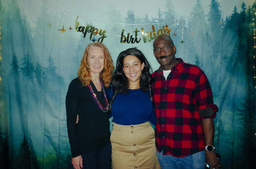Sierra, wearing a khaki skirt, blue top and flowing black wig, stands between her parents. Her mother is dressed as a vampire and her father is dressed in a plaid shirt with a press on moustache. They are posed in front of a photo booth back-drop of a foggy forest with golden Happy Birthday banner above.