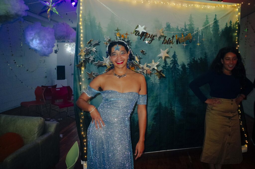 Kyra poses with hand on hip wearing a sparkling floor length off the shoulder gown and a crown of silver stars. Sierra poses in a similar way in a khaki skirt and flowing black wig. Both are posed in front of a photo booth back-drop of a foggy forest with golden Happy Birthday banner above.