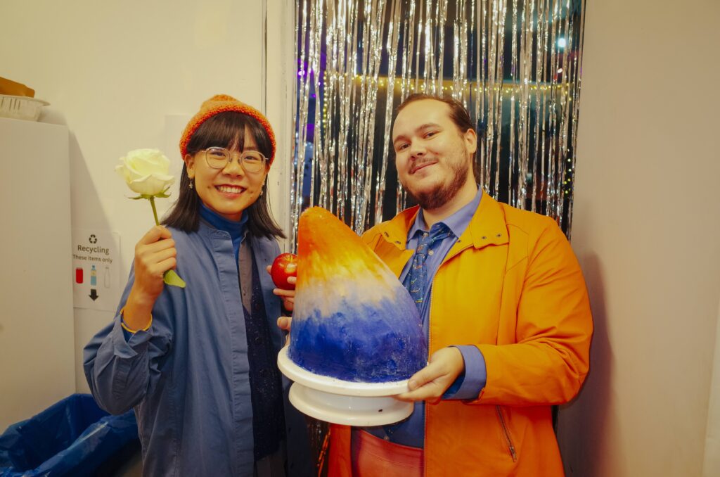 Two people smile widely, one is holding a white rose and an apple and the other is holding a large teardrop shaped cake iced in a vivid ombre in the colours of sunset. Their outfits match the pallet with layers ranging from bright oranges to midnight blues.
